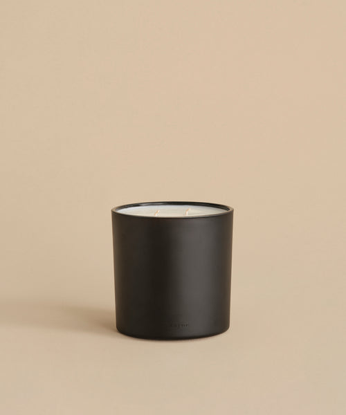 The Large Aspen Glass Candle features two wicks and white wax in a matte black holder. Hand-poured in Los Angeles, it sits on a neutral beige surface with a matching background.