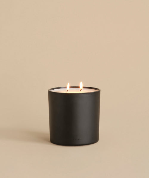 A black matte candle holder displays the Large Cedar Glass Candle (28oz, double wick), glowing warmly on a smooth beige surface against a matching background—an elegant piece inspired by Oprah’s Favorite Things.