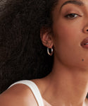 A close-up shows a woman with curly black hair wearing Large Sigrid Hoops in sterling silver and a white top. Only the right side of her face and shoulder are visible, featuring a beauty mark near her lip that highlights the handmade design.