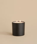 The Large Sun Valley Glass Candle, featuring a hand-poured white two-wick candle in a black matte holder, burns brightly with both flames lit against a plain beige background.