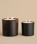 A large, hand-poured Sun Valley Glass Candle with three wicks stands beside a smaller black candle on a neutral beige surface, both in matte containers against a matching background.
