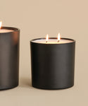 A hand-poured Large Sun Valley Glass Candle with two burning wicks rests in a black matte jar on a neutral surface, beside another similar jar partially shown on the left, all set against a beige background.