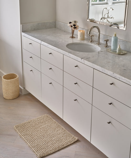 Modern bathroom vanity with white drawers and marble countertop, featuring a round sink, silver faucet, soap dispenser, candle, floral decor, woven trash bin, and the Laurel Bath Mat made from 100% fique on a light wood floor.