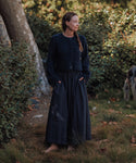 Barefoot on grass, a woman wears the navy Lauren Skirt with a matching jacket, standing in sunlight among green bushes and trees. She faces sideways with hands in her pockets; part of a dog appears on the right.