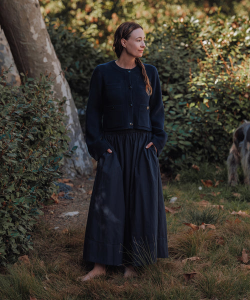 Barefoot on grass, a woman wears the navy Lauren Skirt with a matching jacket, standing in sunlight among green bushes and trees. She faces sideways with hands in her pockets; part of a dog appears on the right.