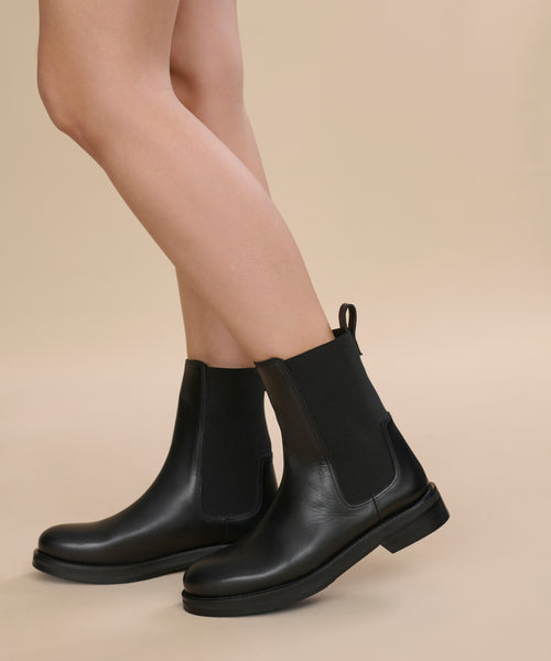 A person wearing the Leather Alden Boot—a black, weather-friendly Chelsea boot with elastic side panels and a heel loop—stands on a beige background, showing only the lower legs and feet.