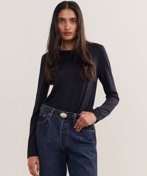A person with long dark hair wears a fitted black top and high-waisted blue jeans styled with the Leather Audrey Belt, featuring a brass buckle, standing against a plain light background.