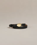 The Leather Audrey Belt, crafted from black premium calf leather with a shiny oval-shaped brass buckle, is coiled on a plain light beige background.