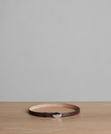 The Leather Audrey Belt, crafted in Italy from brown calf leather, is displayed in a circular shape on a light wooden surface with a gray background.