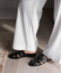 A person wearing white wide-leg pants and black Leather Camilla Mules stands on a light wooden floor beside a beige textured rug.
