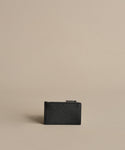The Leather Card Holder, crafted in small black textured leather, stands upright on a beige surface with a minimalist background—perfect for keeping your travel cards organized on any journey.