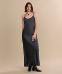 A woman with shoulder-length brown hair and a relaxed expression stands against a beige background, wearing a long dark slip dress, a pendant necklace, and the Leather Carmine Boot.