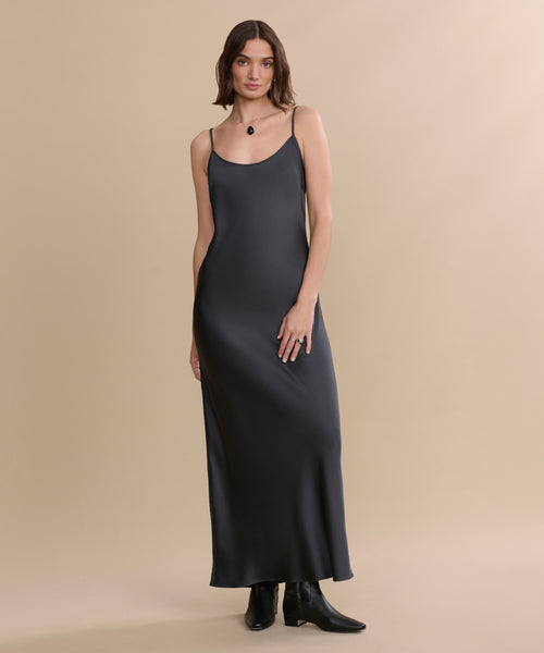 A woman with shoulder-length brown hair and a relaxed expression stands against a beige background, wearing a long dark slip dress, a pendant necklace, and the Leather Carmine Boot.
