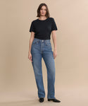A person with shoulder-length brown hair wears a black t-shirt, high-waisted blue jeans, and the Leather Carmine Boot, standing against a plain beige background.