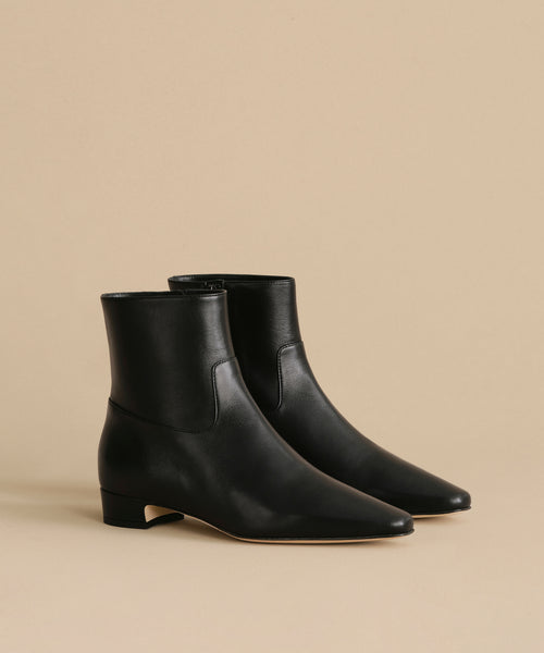 The Leather Carmine Boot, a pair of black leather ankle boots with low heels, pointed toes, and side zippers, showcases a sleek minimalist design against a beige background.