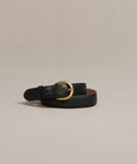 The Classic Belt, crafted from nappa leather in Italy with a gold oval buckle, is displayed coiled on a light beige surface against a neutral background.