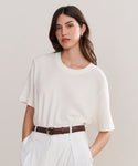 A woman with long brown hair wears a white short-sleeve tee, white high-waisted pants, and the Classic Belt, standing against a plain light background and looking at the camera with a neutral expression.