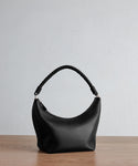 The Crescent Shoulder Bag, crafted in Italy from soft black leather, showcases a curved silhouette and twisted handle as it rests on a wooden surface against a plain light gray background.