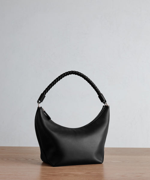 The Crescent Shoulder Bag, crafted in Italy from soft black leather, showcases a curved silhouette and twisted handle as it rests on a wooden surface against a plain light gray background.