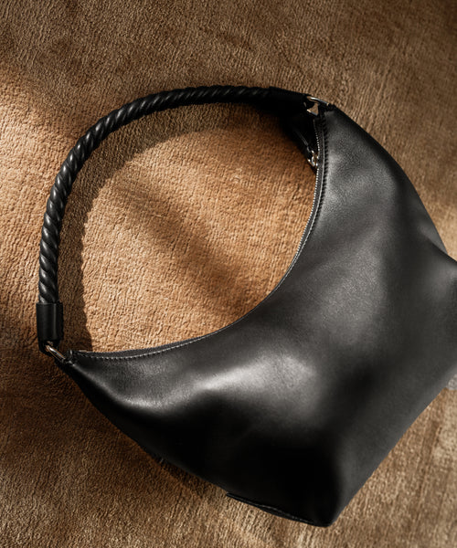 The Crescent Shoulder Bag, crafted by hand in Italy from soft black leather, rests on a tan fabric surface and is illuminated by natural light.