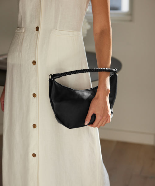 A person in a sleeveless, button-down cream dress holds the Crescent Shoulder Bag, crafted from soft leather and handmade in Italy. The softly lit background features part of a table and a light-colored floor.