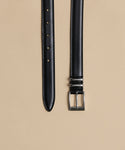The Leather Danner Belt, with a minimalist black leather design and silver rectangular buckle, is shown laid flat on a beige background, displaying punch holes at one end and the buckle with loops at the other.