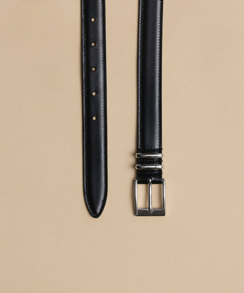 The Leather Danner Belt, with a minimalist black leather design and silver rectangular buckle, is shown laid flat on a beige background, displaying punch holes at one end and the buckle with loops at the other.