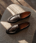 The Leather Dylan Loafer, crafted in Italy from black leather with brown interiors, rests on a textured light surface and is softly illuminated by natural light.