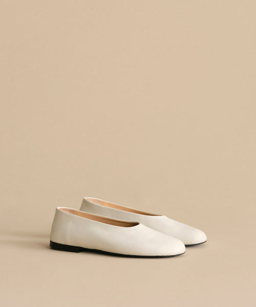 The Leather Elodie Flat, an Italian-made leather shoe with black soles, is displayed on a light beige surface against a matching background, highlighting its timeless and feminine silhouette.