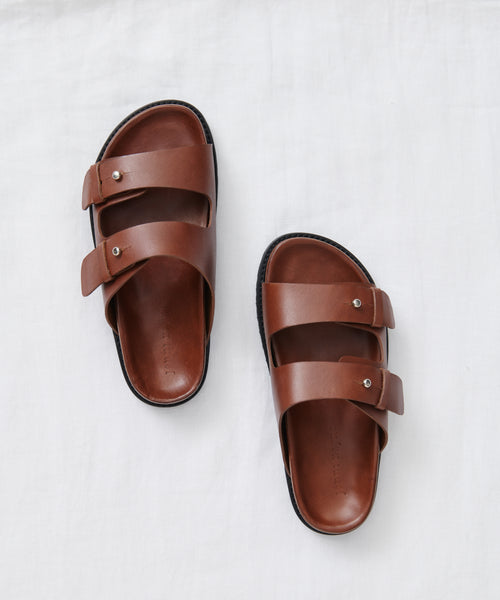 The Hutton Slide Sandal, featuring two leather straps with metal buckles and crafted in Portugal, is shown on a white fabric background.