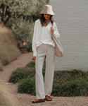 A woman stands outdoors by a white brick wall, surrounded by greenery. She wears a beige bucket hat, loose white sweater, light linen trousers, and the Hutton Slide Sandal from Portugal, carrying a beige shoulder bag.