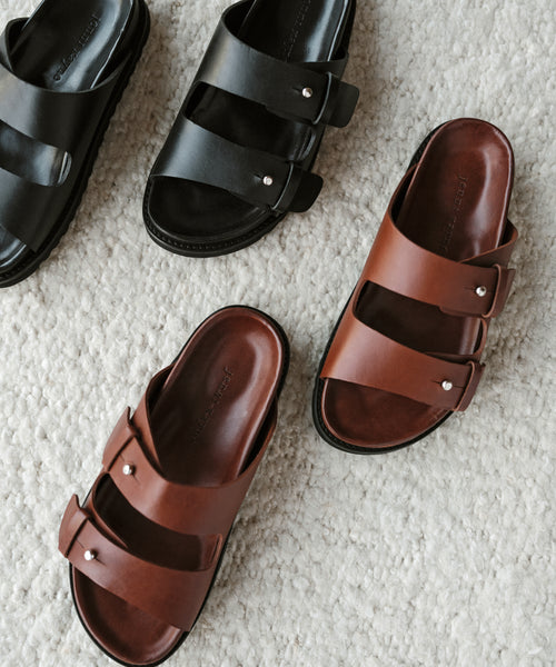 Two pairs of Hutton Slide Sandal, one black and one brown with double straps and silver buckles, made in Portugal, rest on a light, textured carpet.