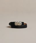 The Leather Jules Belt, a black leather belt with a large rounded rope-twist vintage silver buckle, is coiled and displayed on a neutral beige background.