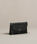 The Loren Clutch Bag is a small black Italian-made accessory with a curved flap, silver button clasp, and thin shoulder strap, shown on a simple beige background.