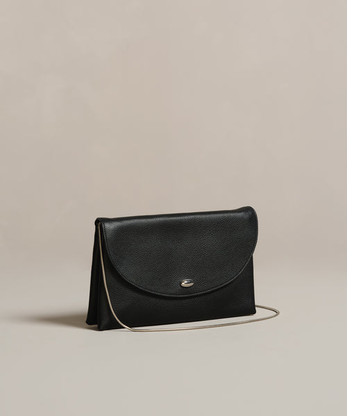 The Loren Clutch Bag is a small black Italian-made accessory with a curved flap, silver button clasp, and thin shoulder strap, shown on a simple beige background.