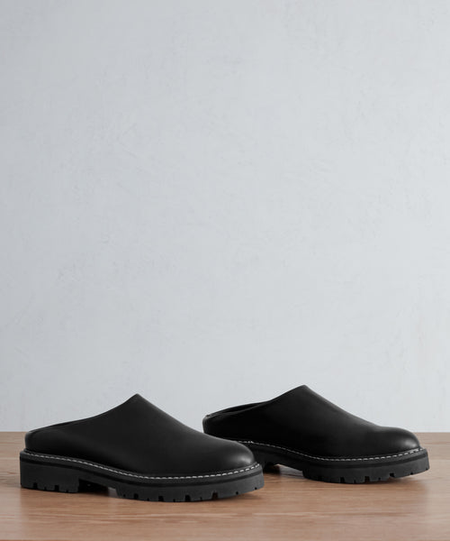 A pair of Leather Lug Mule shoes with thick, rugged soles sit side by side on a wooden surface against a plain, light background.