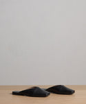 The Mia Mule, a pair of black Italian leather square-toe slip-on flats, is placed side by side on a light wooden surface with a plain light gray background.