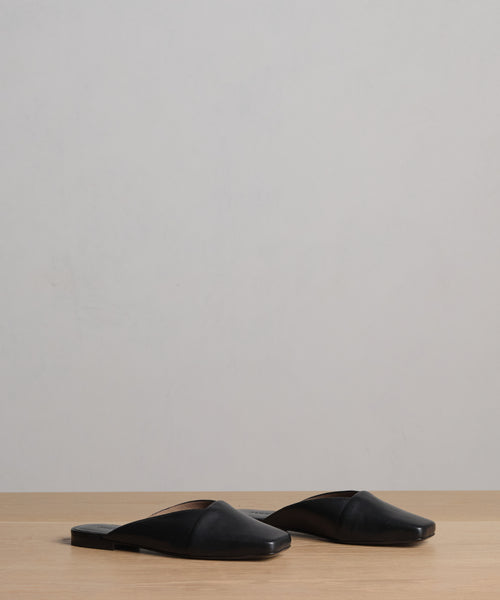 The Mia Mule, a pair of black Italian leather square-toe slip-on flats, is placed side by side on a light wooden surface with a plain light gray background.