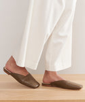 A person stands on a light wooden surface wearing loose white pants and the Mia Mule brown Italian leather shoes, with only their lower legs and feet visible.