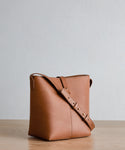 The Mini Leather Bucket Bag, crafted in Italy from tan vachetta leather with a wide adjustable strap, is displayed on a light wooden surface against a neutral textured background.
