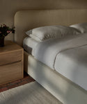 A bed with a white Dusk Linen Duvet Cover and two pillows sits by a wooden nightstand topped with a vase of dark flowers, all arranged on a cream textured rug over a brick floor.
