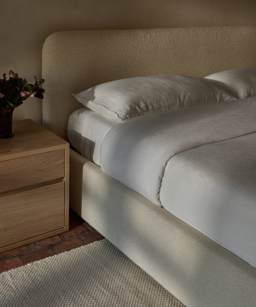 A bed with a white Dusk Linen Duvet Cover and two pillows sits by a wooden nightstand topped with a vase of dark flowers, all arranged on a cream textured rug over a brick floor.