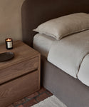 A neatly made bed with beige lightweight linen bedding, featuring the Dusk Linen Fitted Sheet made in Portugal, sits beside a wooden nightstand with a lit candle and dark dish on a textured brick rug floor.
