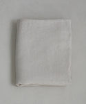 The Dusk Linen Flat Sheet, in light beige natural linen, is neatly folded and lies flat on a textured pale surface, highlighting the understated elegance of luxury bedding.