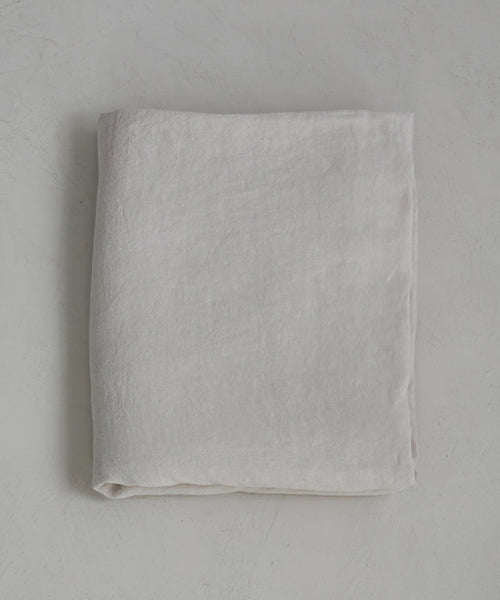 The Dusk Linen Flat Sheet, in light beige natural linen, is neatly folded and lies flat on a textured pale surface, highlighting the understated elegance of luxury bedding.