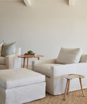 A minimal, neutral-toned living room with a white armchair, the Harbor Ottoman in natural linen handmade in Los Angeles, a rustic wooden stool, round side table with books and plant, and a beige textured rug on the floor.