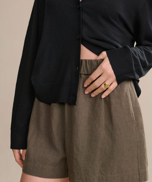 A person wears a black long-sleeve top and the Linen Logan Short in dark moss, resting their left hand with a gold ring on their waist. Their face is hidden, highlighting an effortlessly stylish warm-weather look with the Linen Logan Short.