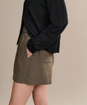 Standing sideways with one hand in the pocket of their dark moss Linen Logan Short and wearing a black long-sleeve top, this look is perfect for warm-weather days against a neutral background.