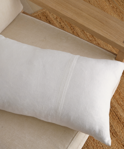 A Linen Lumbar Pillow with a feather down insert sits on a beige cushioned chair with wooden armrests, set atop a textured brown rug.