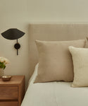 A neatly made bed with a Linen Pillow and light blanket sits beside a wooden nightstand holding a small bowl, white flowers in a vase, and a modern black wall sconce, giving the room a calm, minimalist style.